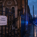 Beschädigtes Metalltor vor einem ehemaligen Synagogengebäude in einer Londoner Straße bei Dämmerung, mit sichtbaren Brandspuren am Schloss und Blaulicht eines Polizeifahrzeugs im Hintergrund.