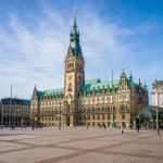 Das Hamburger Rathaus erhebt sich im warmen Tageslicht über dem weitläufigen Rathausmarkt. Der historische Sandsteinbau mit seinem markanten Uhrturm und den grünen Kupferdächern steht im Mittelpunkt des Bildes. Auf dem Platz sind vereinzelt Passanten unterwegs, während der blaue Himmel und die offene Atmosphäre die Bedeutung des Rathausmarktes als zentralen Ort des öffentlichen Lebens in Hamburg unterstreichen.