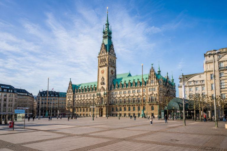Das Hamburger Rathaus erhebt sich im warmen Tageslicht über dem weitläufigen Rathausmarkt. Der historische Sandsteinbau mit seinem markanten Uhrturm und den grünen Kupferdächern steht im Mittelpunkt des Bildes. Auf dem Platz sind vereinzelt Passanten unterwegs, während der blaue Himmel und die offene Atmosphäre die Bedeutung des Rathausmarktes als zentralen Ort des öffentlichen Lebens in Hamburg unterstreichen.