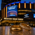 Zwei goldene Trauringe liegen im Vordergrund vor dem beleuchteten Madison Square Garden in New York. Die Arena erstrahlt in den Abendstunden, während die Ringe symbolisch für die Hochzeit von Taylor Swift und Travis Kelce stehen.