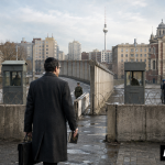 Ein jüdischer Kantor mit Kippa und Gebetbuch steht vor einem Grenzübergang der Berliner Mauer. Auf beiden Seiten sind Synagogen und Gemeindemitglieder zu sehen. Unten rechts steht „AI GENERATED“.