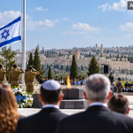Gedenkzeremonie in Jerusalem bei Tageslicht mit israelischer Flagge auf halbmast, salutierenden Soldaten, Kränzen und Blick auf die Altstadt.